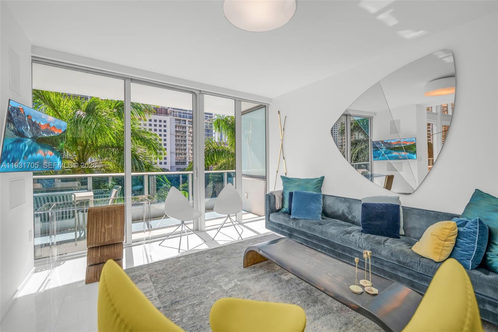 A modern living room features a blue velvet sofa, yellow chairs, and a large mirror — 200 Biscayne Boulevard Way # 309, Miami FL 33131, MIAMI, FL
