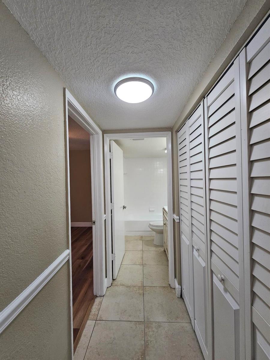 A hallway with tiled flooring leads to a bathroom, featuring louvered closet doors on the right — 1541 Windorah Way # C, West Palm Beach FL 33411, WEST PALM BEACH, FL