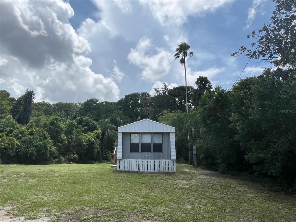 A small, single-story manufactured home sits on a grassy lot surrounded by dense trees — 9860 GOLDEN LOOP, NEW PORT RICHEY, FL