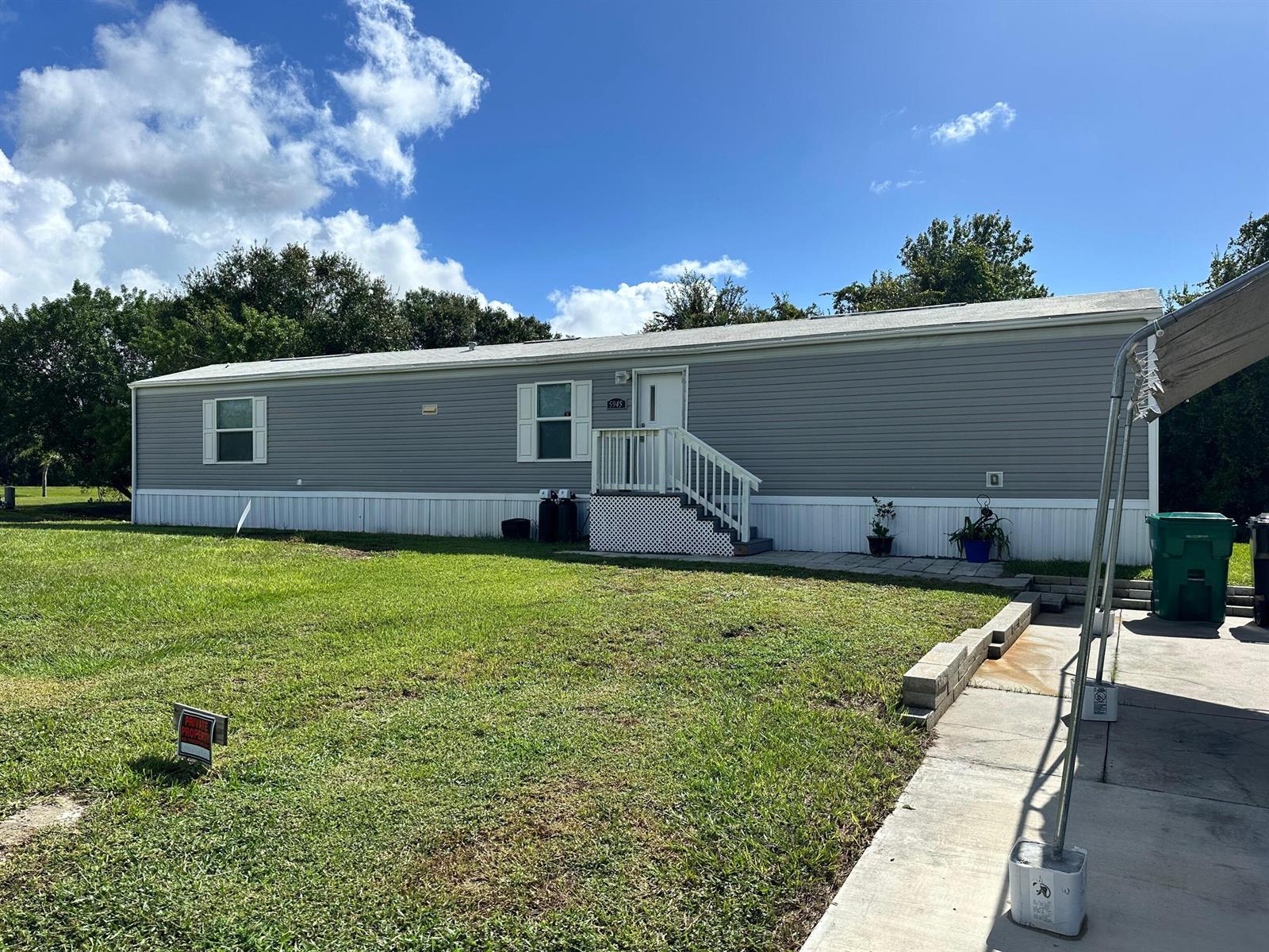 A single-story manufactured home with grey siding features a white front door and stairs — 5945 SE 95th Trail Trail, Okeechobee FL 34974, OKEECHOBEE, FL