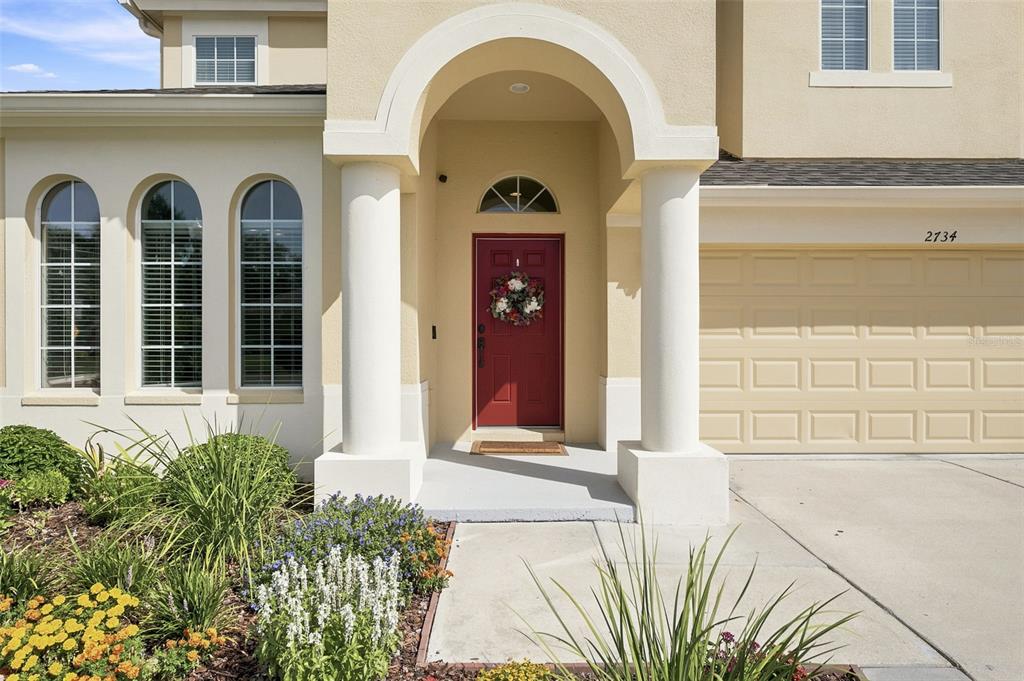 A beige house exterior features a red front door, arched windows, and a two-car garage — 2734 SILVERMOSS DRIVE, WESLEY CHAPEL, FL