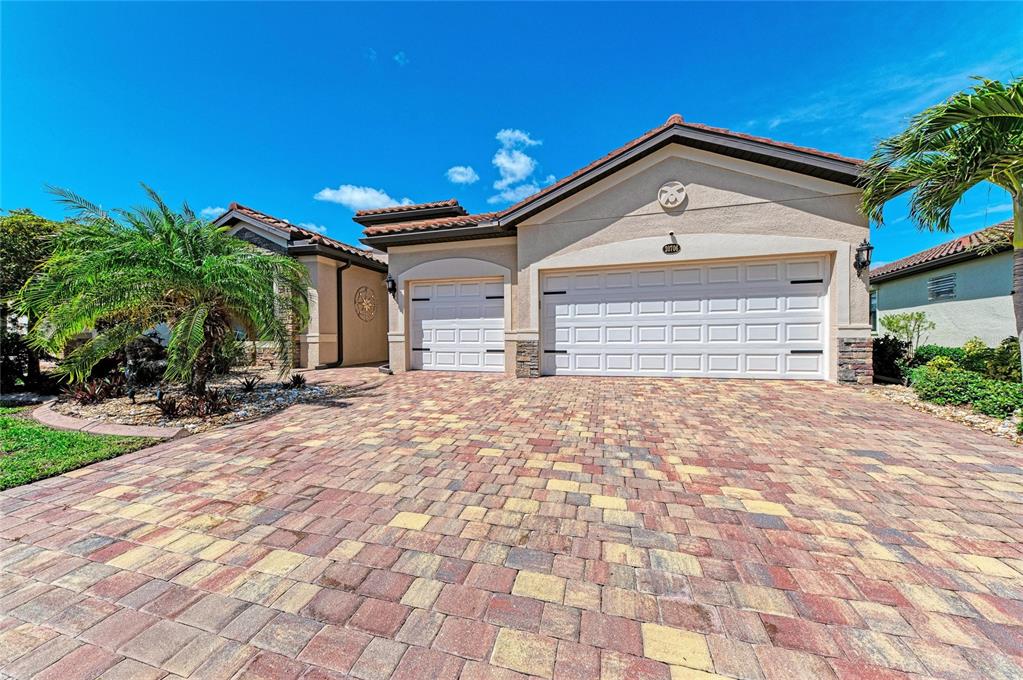 A beige house features a multi-car garage and a patterned paver driveway under a blue sky — 20706 BENISSIMO DRIVE, VENICE, FL