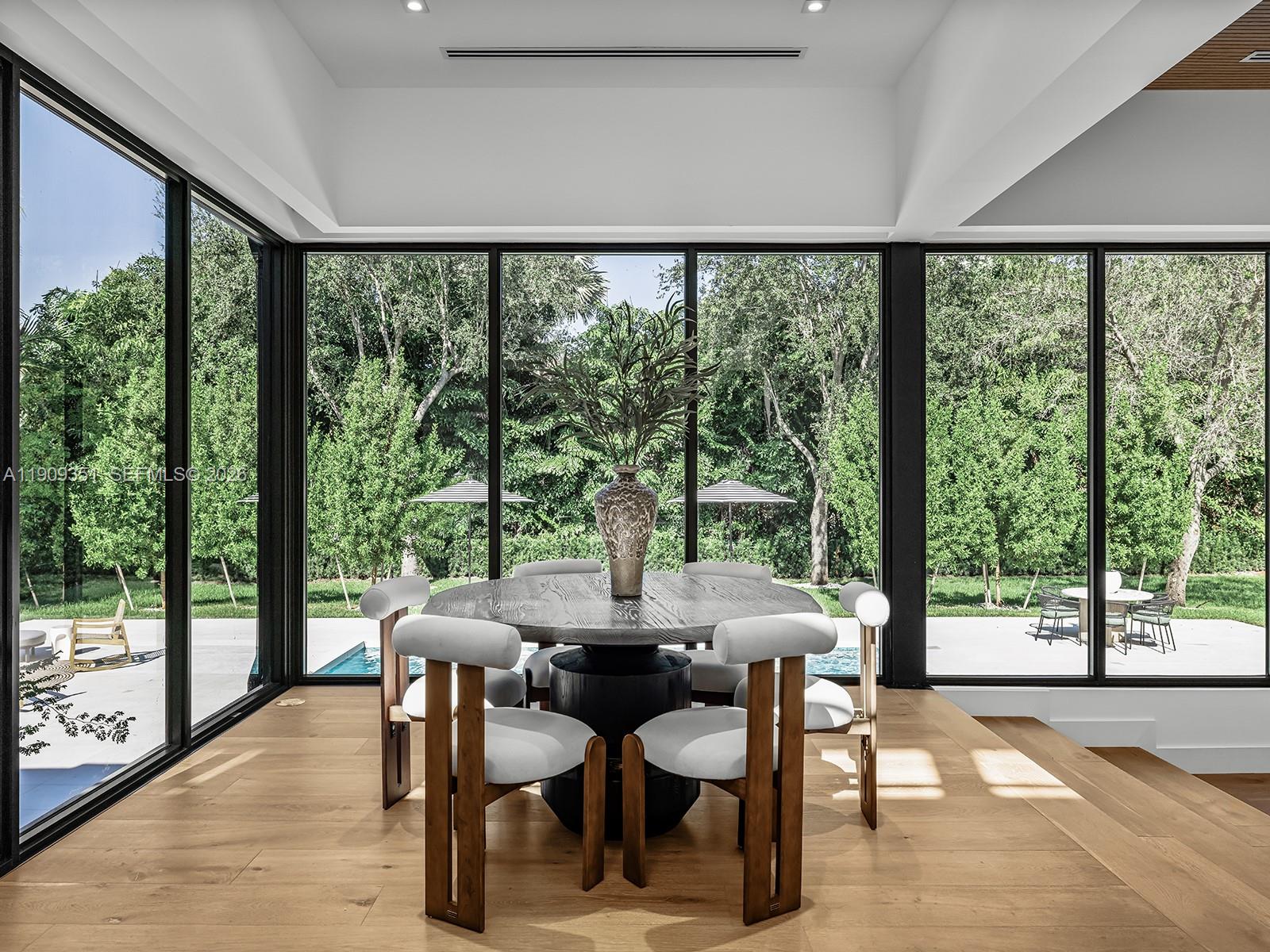 A dining area with a round table and chairs sits before floor-to-ceiling windows overlooking greenery — 7910 Los Pinos Cir, Coral Gables FL 33143, CORAL GABLES, FL