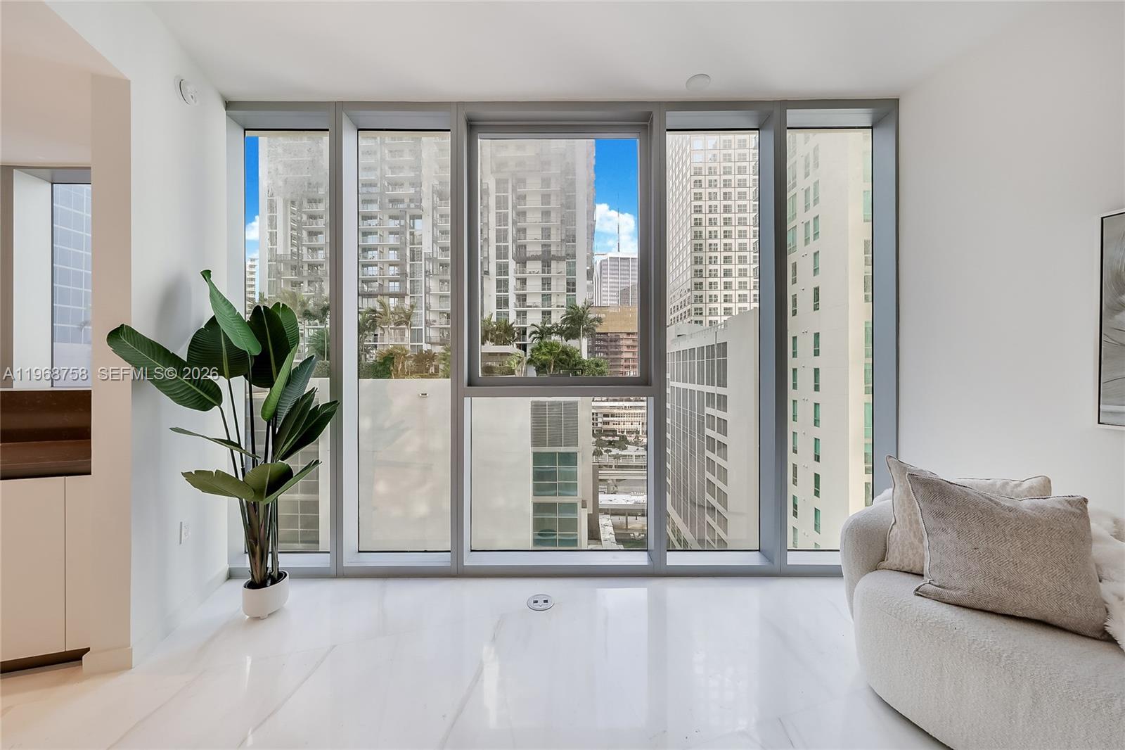 A bright room features floor-to-ceiling windows overlooking city buildings, a potted plant, and marble flooring — 300 Biscayne Boulevard Way # 1105C, Miami FL 33131, MIAMI, FL