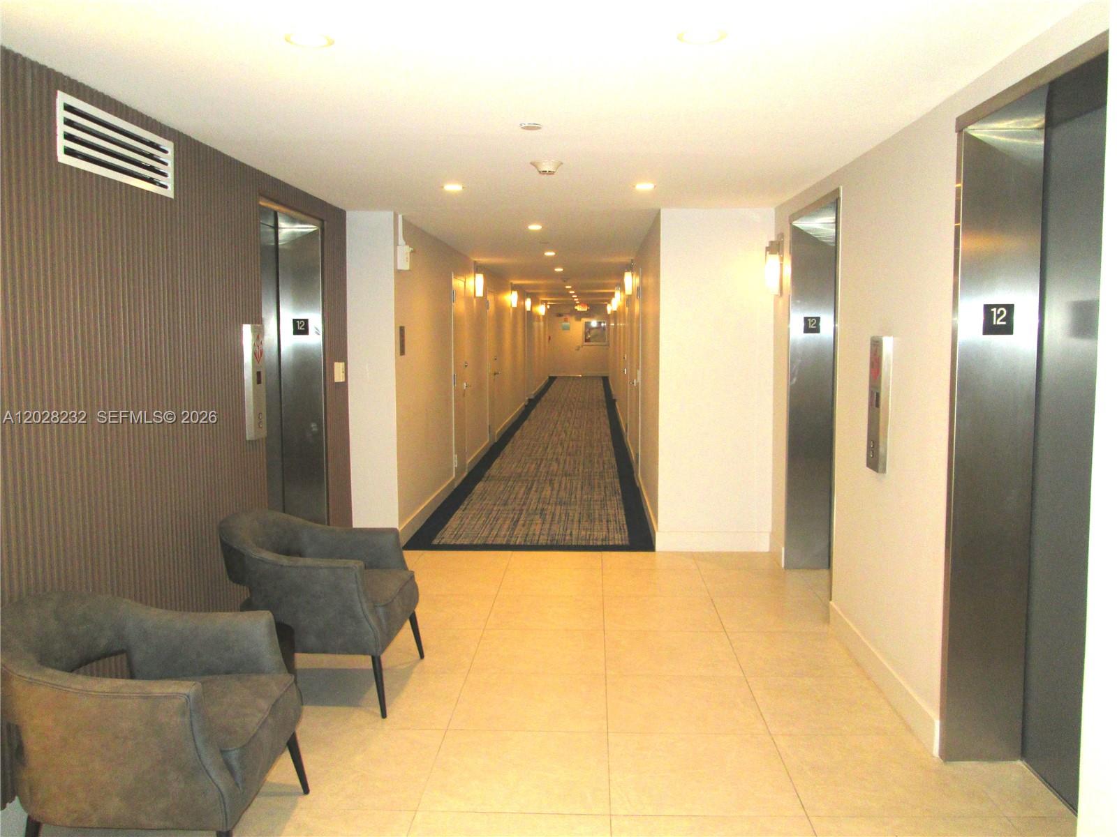 A hallway features two elevators, two gray armchairs, and a long carpeted corridor — 1370 S Ocean Blvd # 1206, Pompano Beach FL 33062, POMPANO BEACH, FL