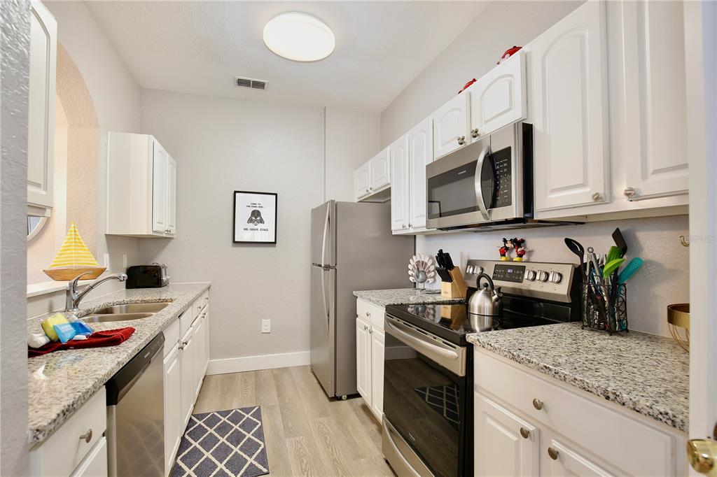 A galley kitchen features white cabinets, granite countertops, stainless steel appliances, and light wood flooring — 2821 ALMATON LOOP UNIT 301, KISSIMMEE, FL
