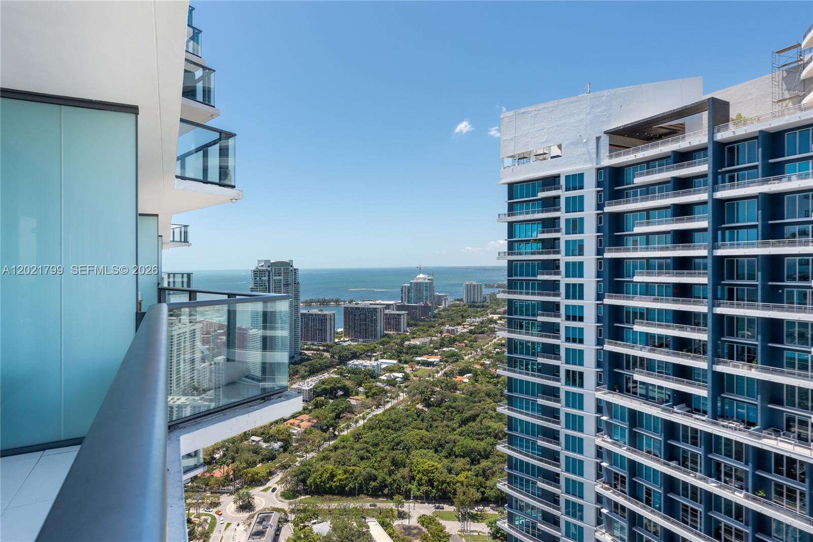 A high-rise balcony overlooks a dense urban landscape, lush green trees, and the distant ocean — 1300 S Miami Ave # 4709, Miami FL 33130, MIAMI, FL