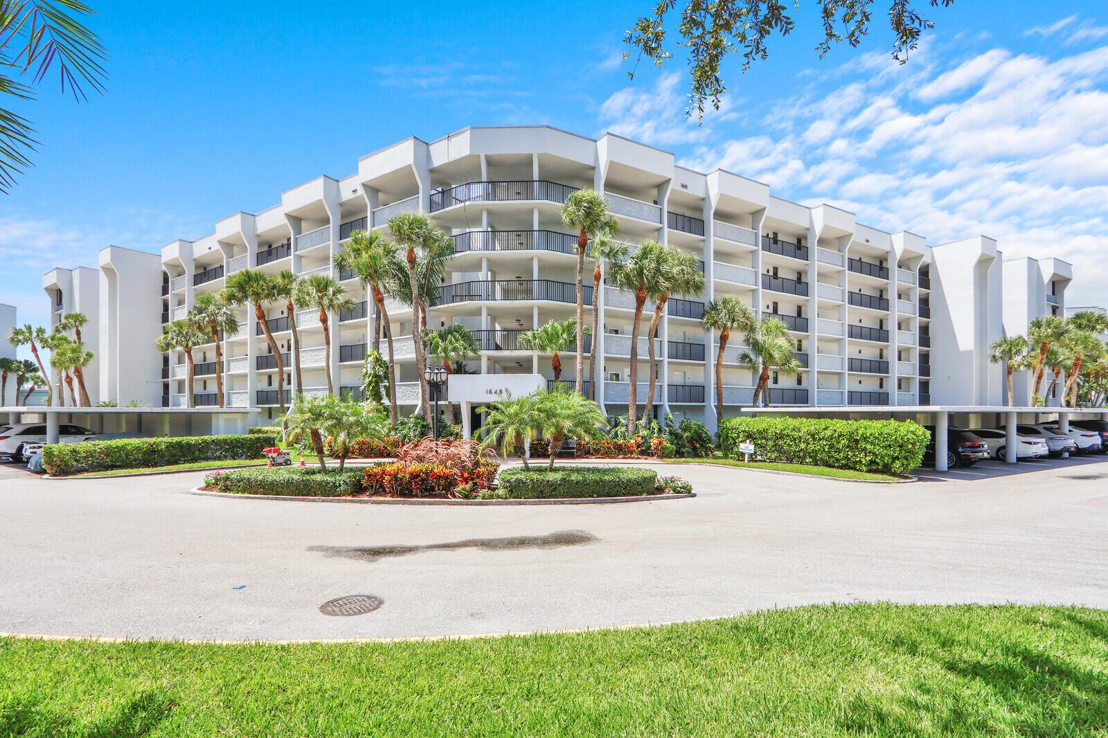 A multi-story white apartment building features balconies and a circular driveway with palm trees — 1648 Jupiter Cove Drive # 214, Jupiter FL 33469, JUPITER, FL