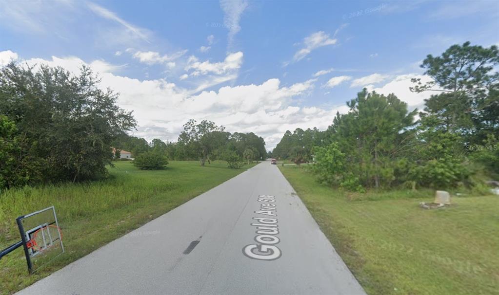 A paved road stretches through a grassy, wooded residential area under a cloudy blue sky — 1756 GOULD AVENUE SW, PALM BAY, FL