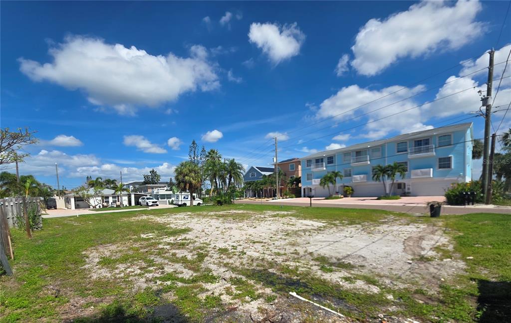 A vacant, sandy lot sits in front of a multi-story blue building under a cloudy sky — 14268 N BAYSHORE DRIVE, MADEIRA BEACH, FL