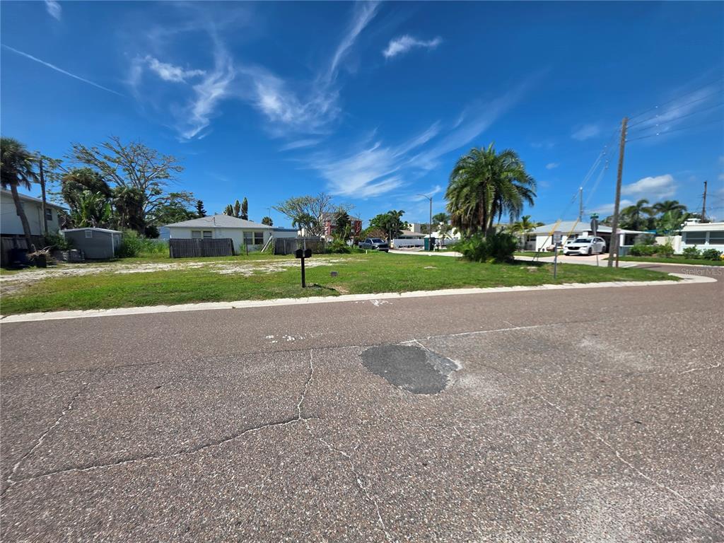 A vacant, grassy residential lot sits along a paved street under a blue sky — 14268 N BAYSHORE DRIVE, MADEIRA BEACH, FL