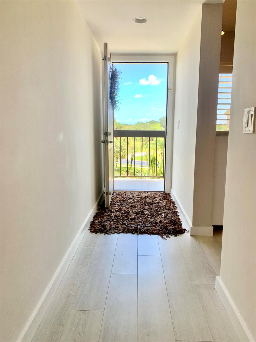 A hallway with light wood-look flooring leads to an open door revealing a balcony view — 1401 Tidal Pointe Boulevard # 406, Jupiter FL 33477, JUPITER, FL