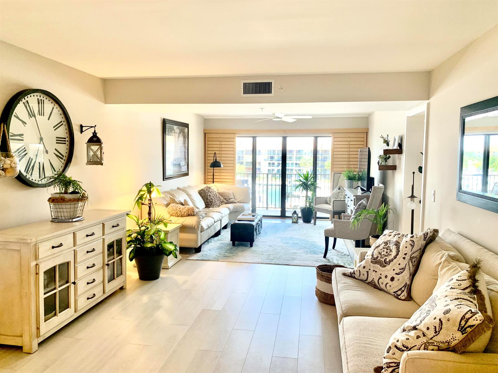 A bright living room features light wood flooring, two sofas, a large wall clock, and balcony access — 1401 Tidal Pointe Boulevard # 406, Jupiter FL 33477, JUPITER, FL