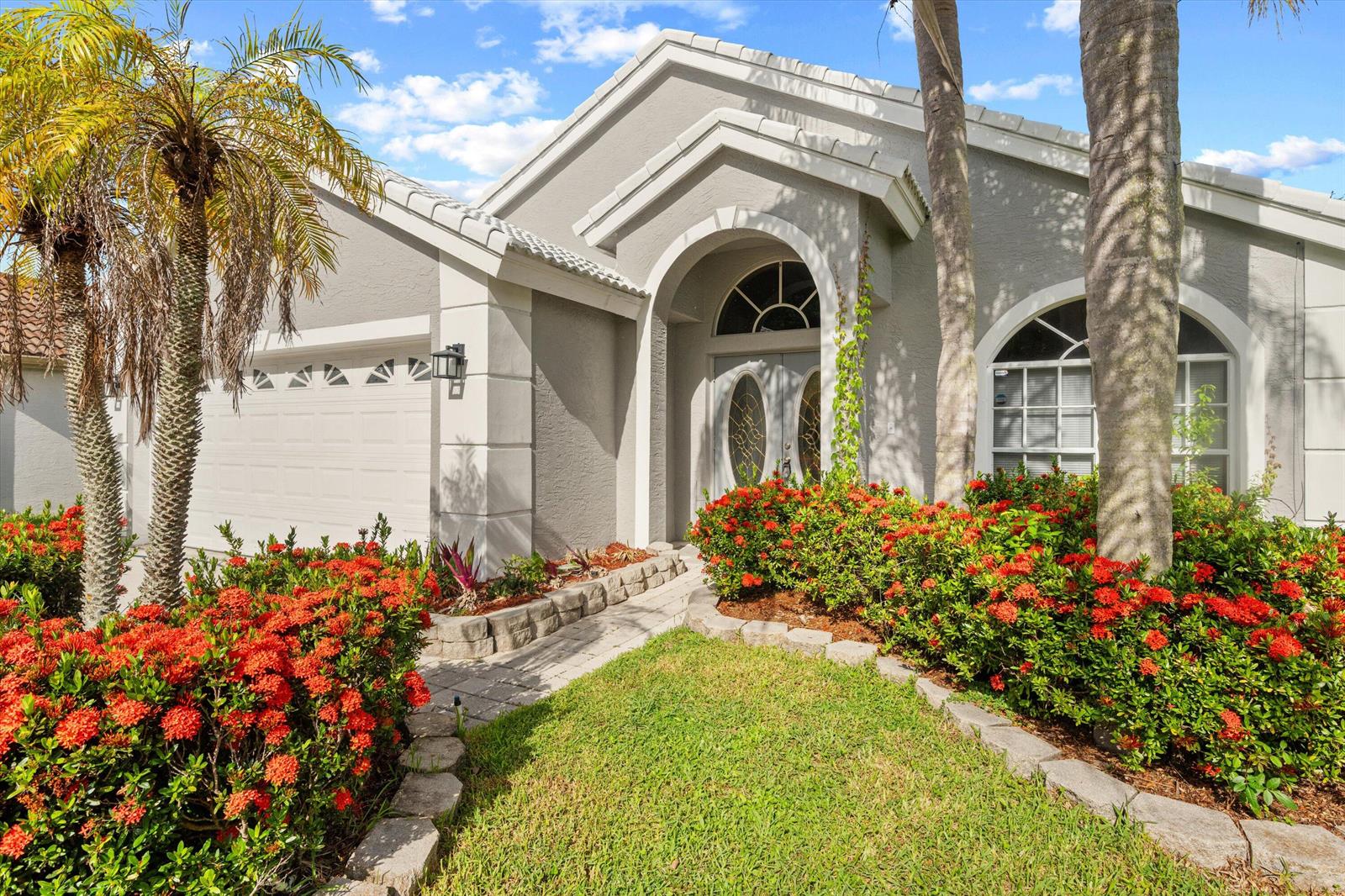 A gray single-story house features a arched entryway, a two-car garage, and red flowering shrubs — 1851 Oak Berry Circle, Wellington FL 33414, WELLINGTON, FL