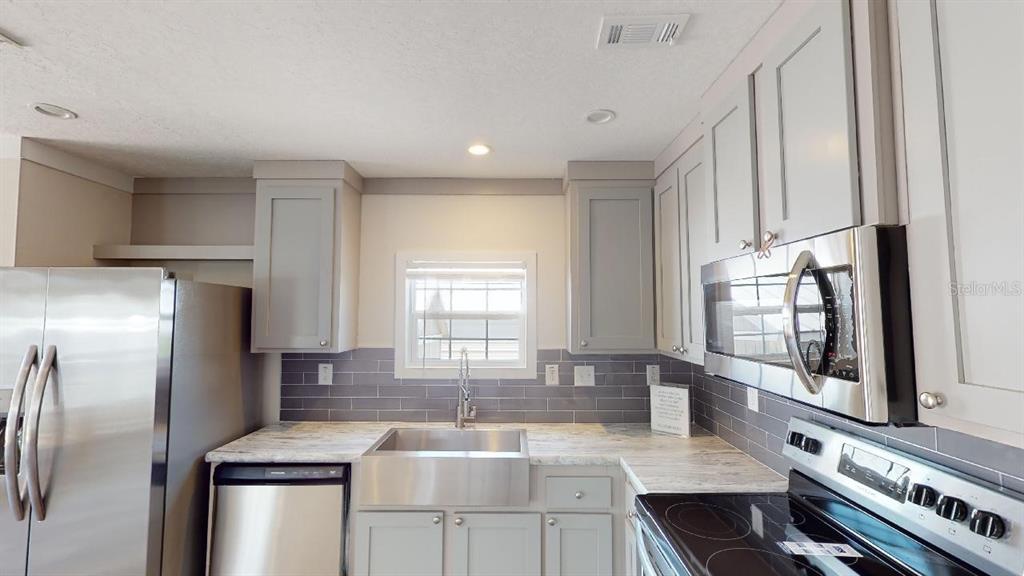 A kitchen features gray cabinets, a farmhouse sink, stainless steel appliances, and a tiled backsplash — 1556 BLUE HERON DRIVE, ENGLEWOOD, FL