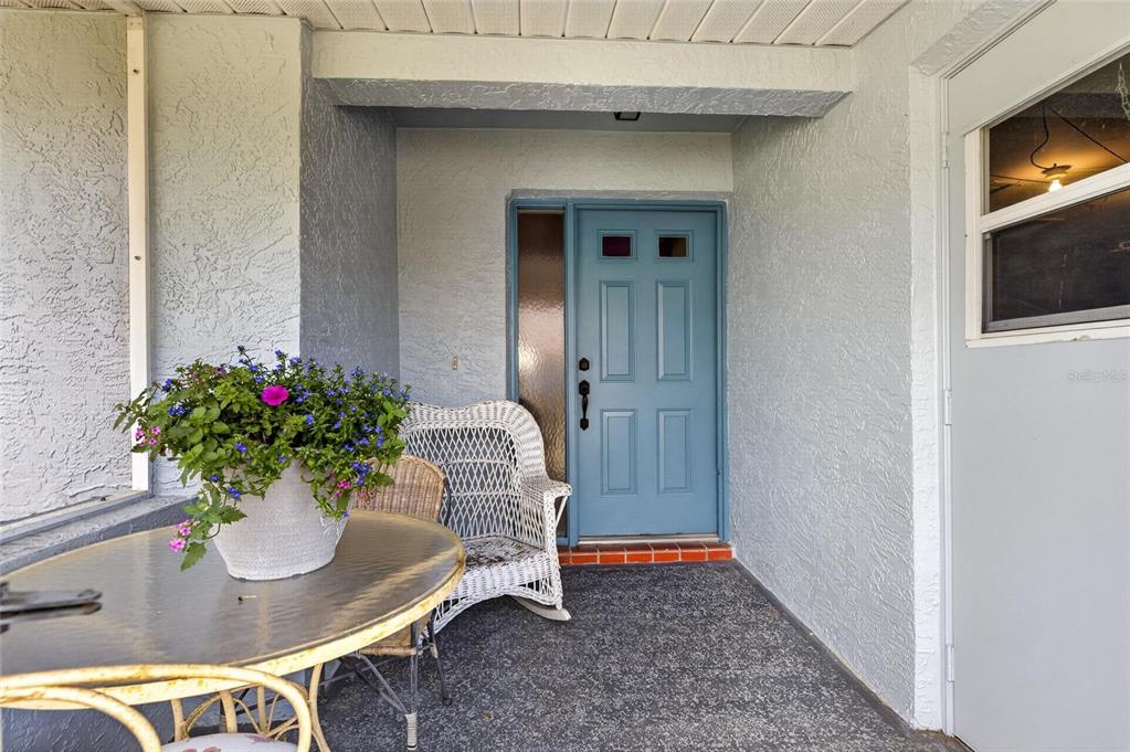 A covered porch features a blue front door, a glass-topped table, and a wicker chair — 1517 INGRAM DRIVE, SUN CITY CENTER, FL