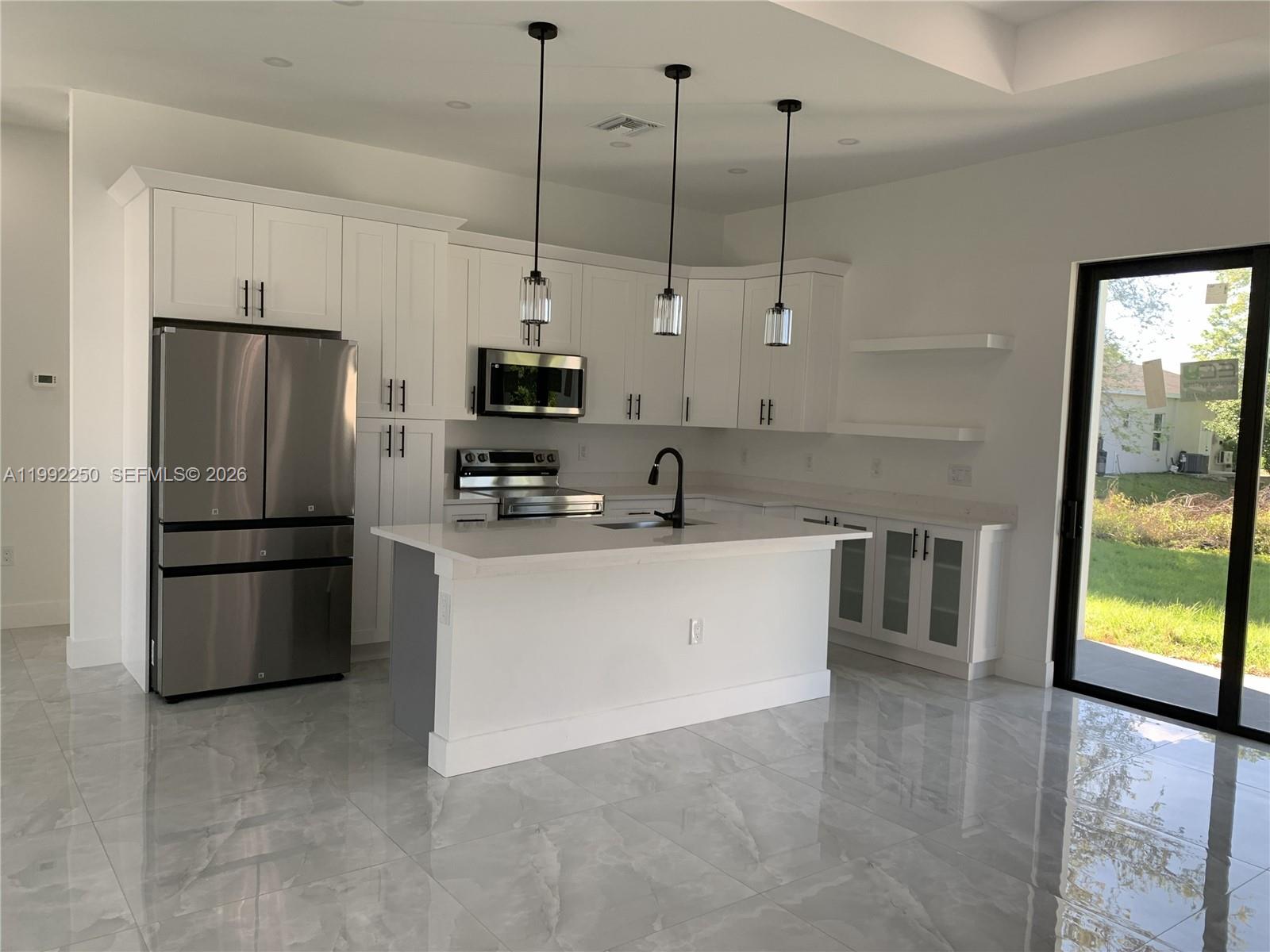 A modern white kitchen features a stainless steel refrigerator, a center island, and tile flooring — 442 Redwing St, Lehigh Acres FL 33974, LEHIGH ACRES, FL