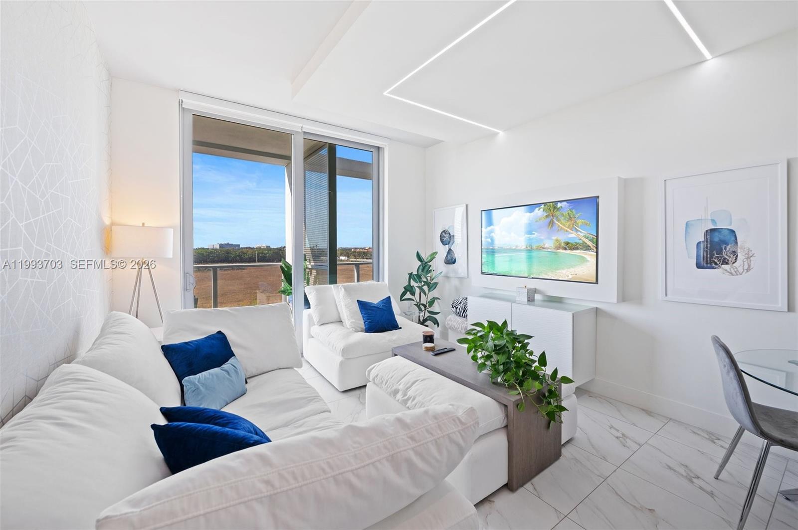 A modern living room features white furniture, blue accent pillows, marble floors, and a balcony — 2000 Metropica Way # 508, Sunrise FL 33323, SUNRISE, FL
