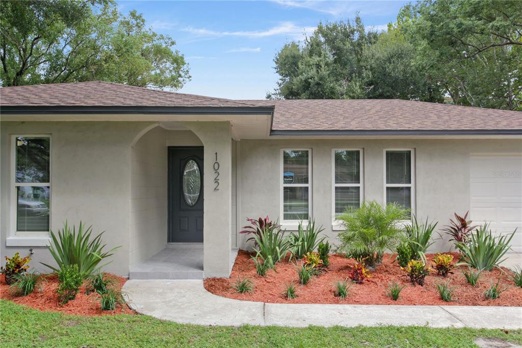 The exterior of a single-story house features a gray stucco facade, a dark door, and landscaping — 1022 NORWOOD DRIVE, DELTONA, FL