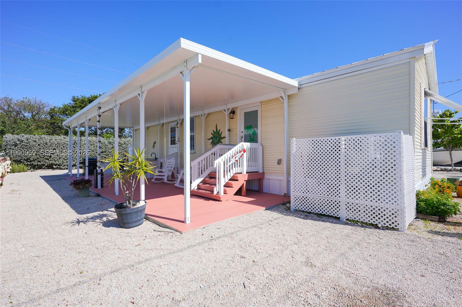 A light yellow mobile home features a covered porch with white railings and gravel landscaping — 112 Tweedy Pie Ter, Key Largo FL 33037, KEY LARGO, FL