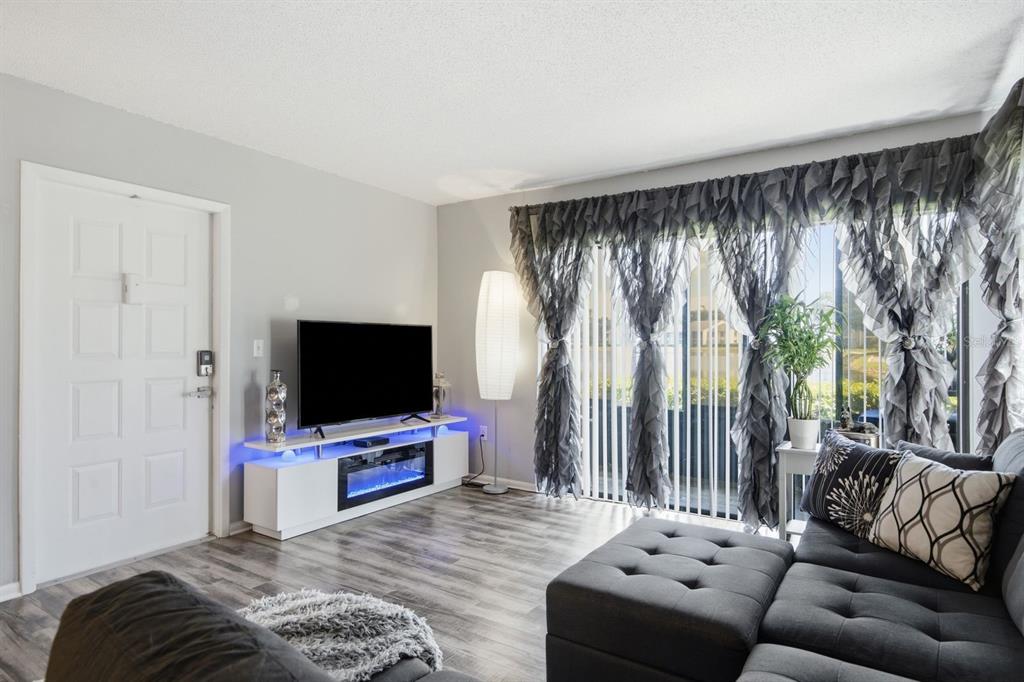 A living room features gray wood-look flooring, a white media console with a fireplace, and curtains — 1179 PINE RIDGE CIRCLE W UNIT G1, TARPON SPRINGS, FL