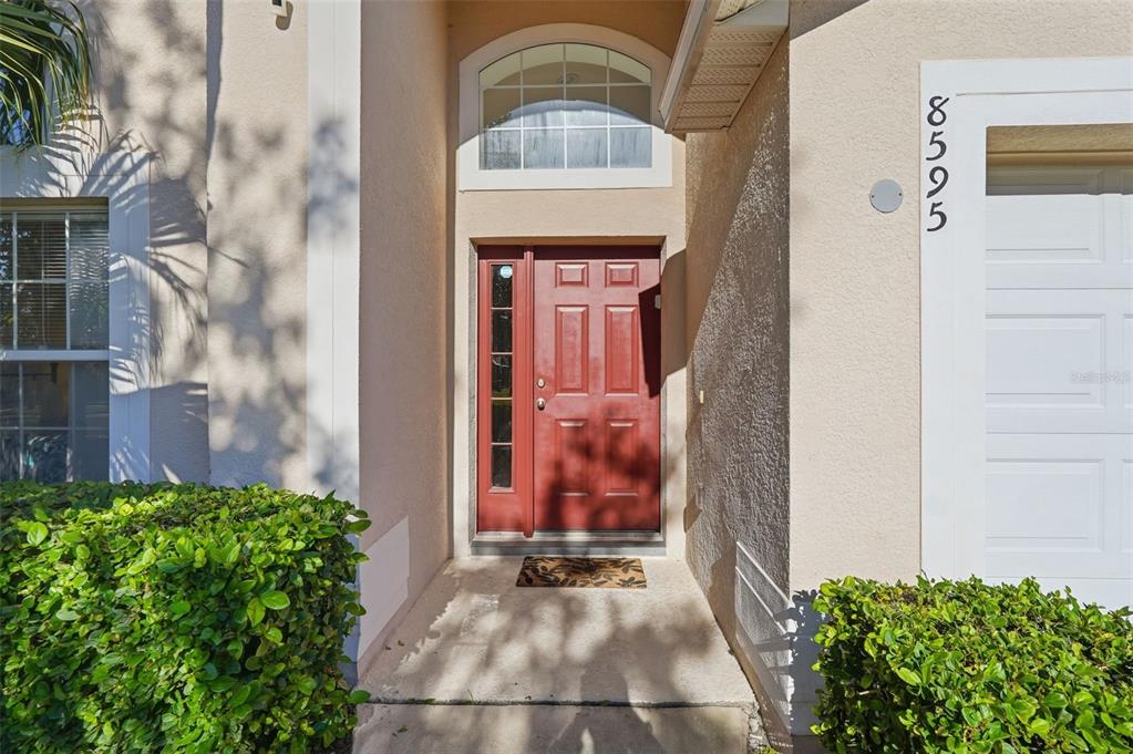 A red front door with a side window and arched transom sits under a porch — 8595 SUNRISE KEY DRIVE, KISSIMMEE, FL