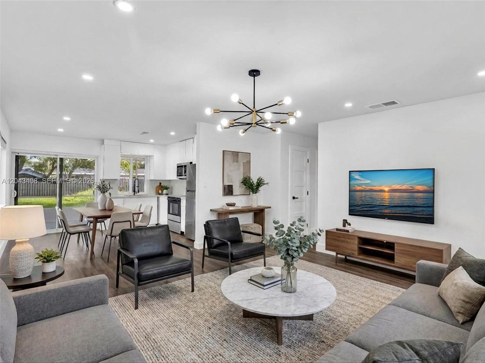 An open-concept living and dining area features a modern chandelier, neutral furniture, and wood flooring — 8145 Sunset Strip, Sunrise FL 33322, SUNRISE, FL