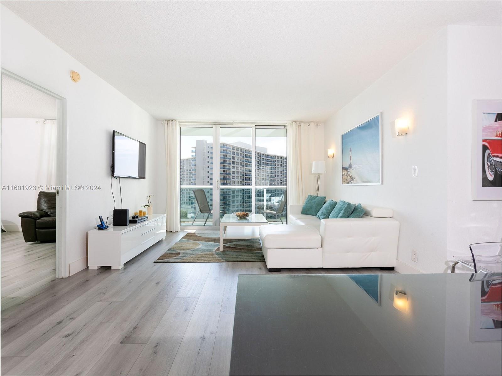A living room features white furniture, a sectional sofa, wood-look flooring, and a balcony view — 3901 S Ocean Dr # 10P, Hollywood FL 33019, HOLLYWOOD, FL
