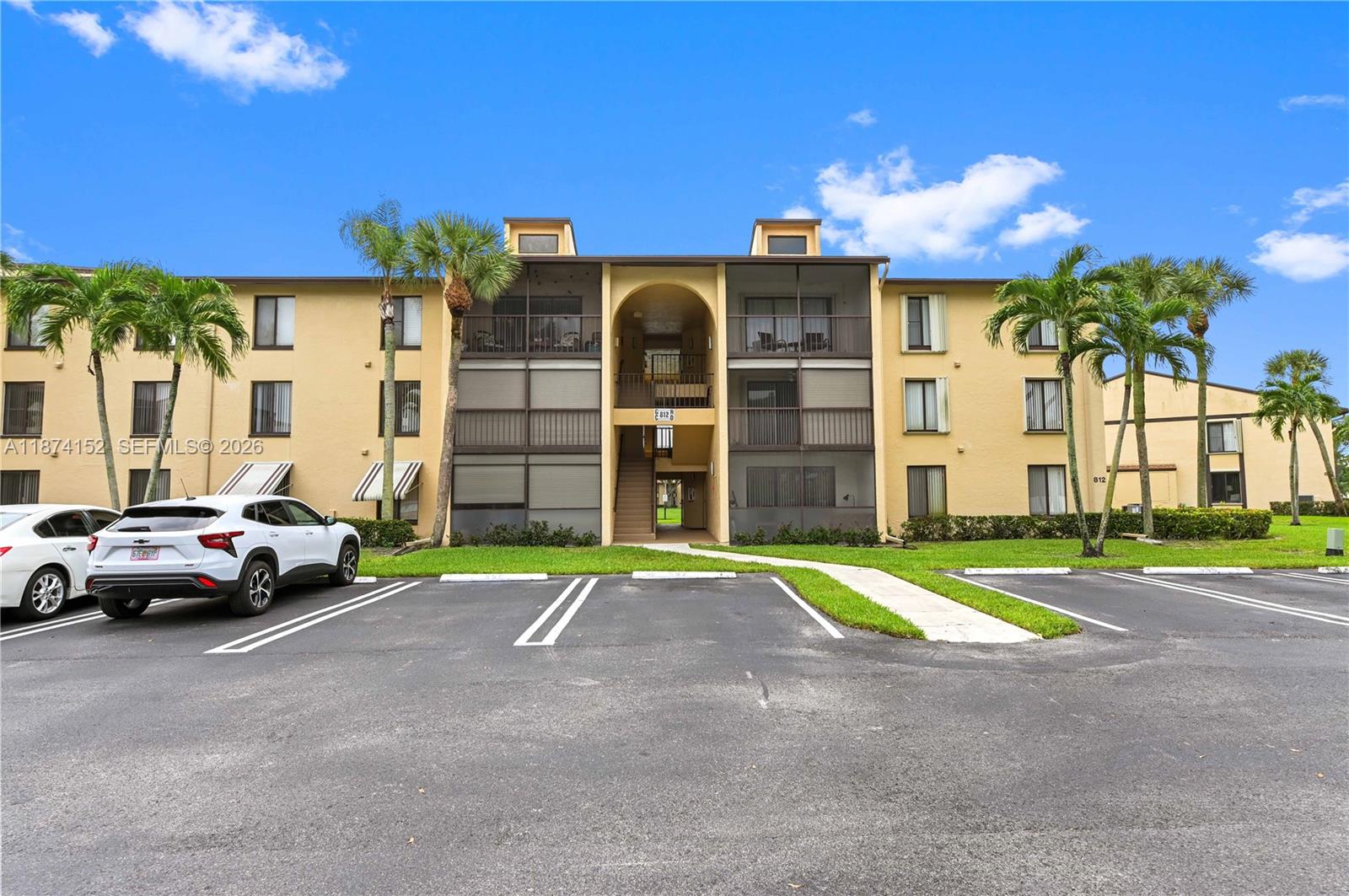 A three-story apartment building with tan stucco walls and screened balconies sits behind a parking lot — 812 Sky Pine Way # C2, Green Acres FL 33415, GREEN ACRES, FL