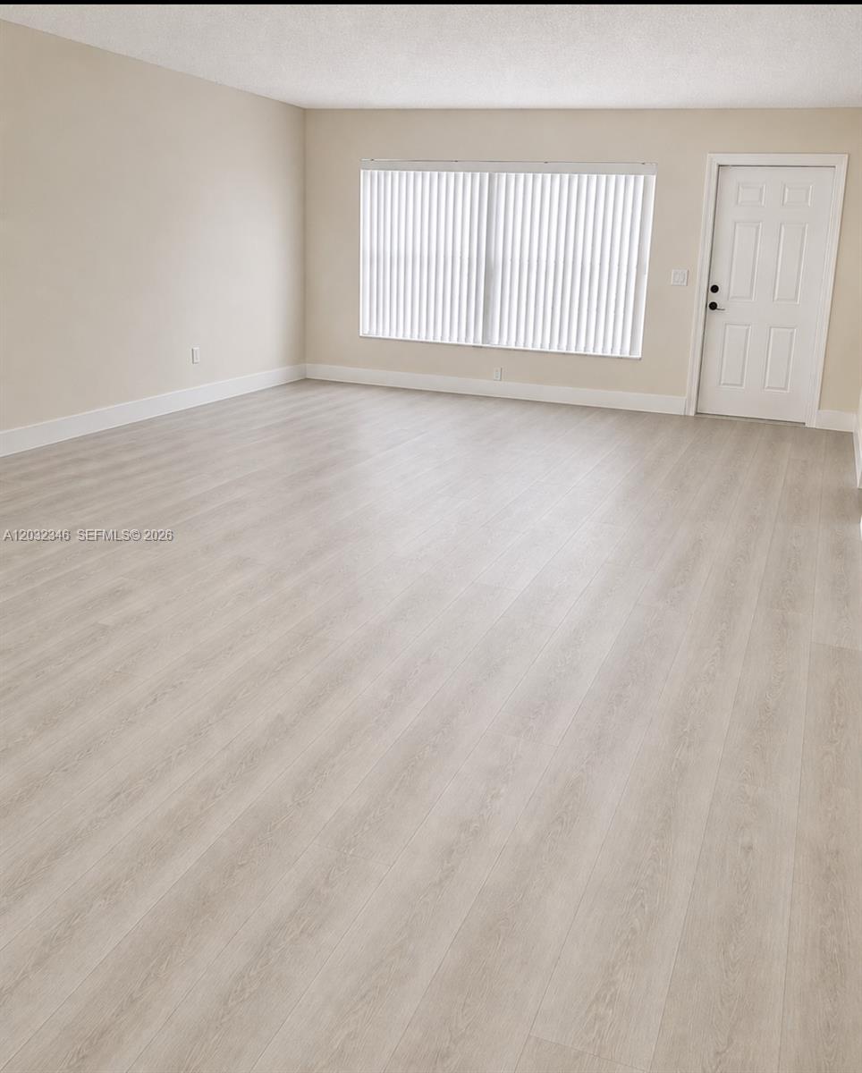 An empty room features light-colored wood-look flooring, beige walls, vertical blinds, and a white door — 22745 SW 65th Way # 0, Boca Raton FL 33428, BOCA RATON, FL