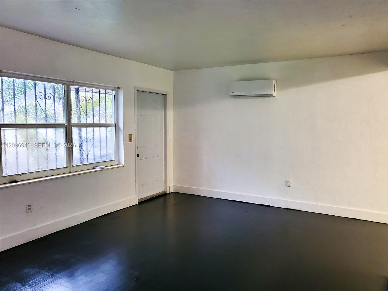 An empty room features white walls, dark flooring, a window with security bars, and an AC unit — 1225 NW 127th St, North Miami FL 33167, NORTH MIAMI, FL