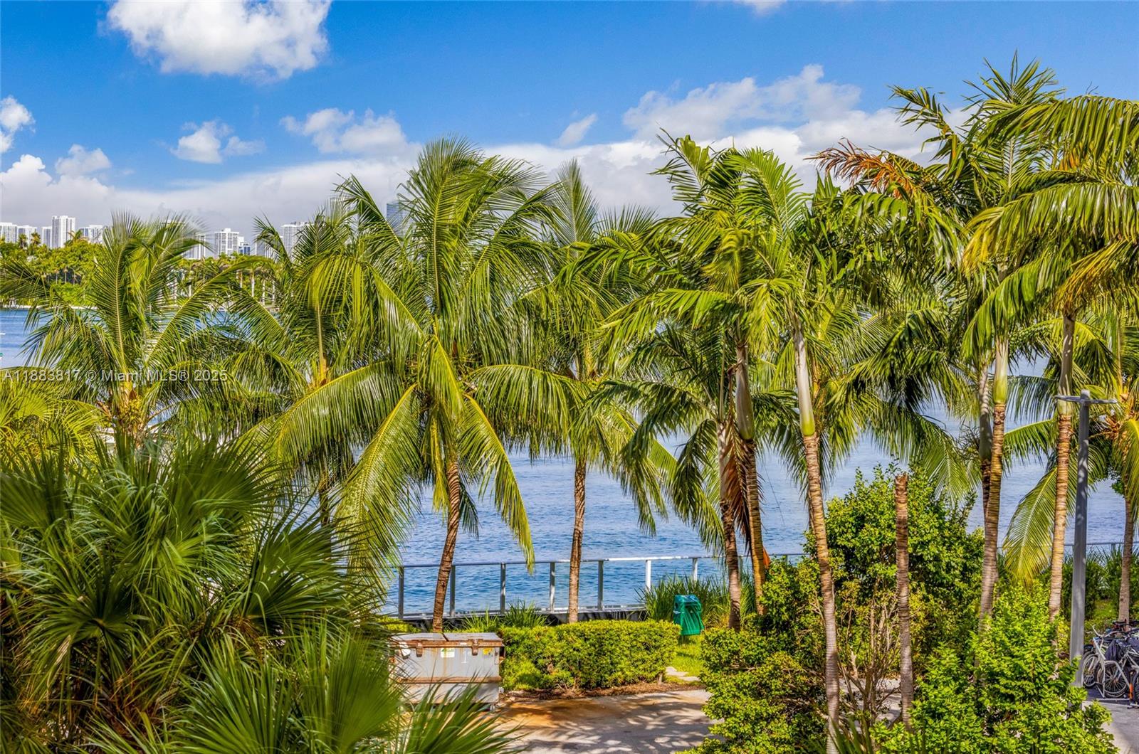 A view of a waterfront area with palm trees, a metal railing, and distant buildings — 800 West Ave # 338, Miami Beach FL 33139, MIAMI BEACH, FL