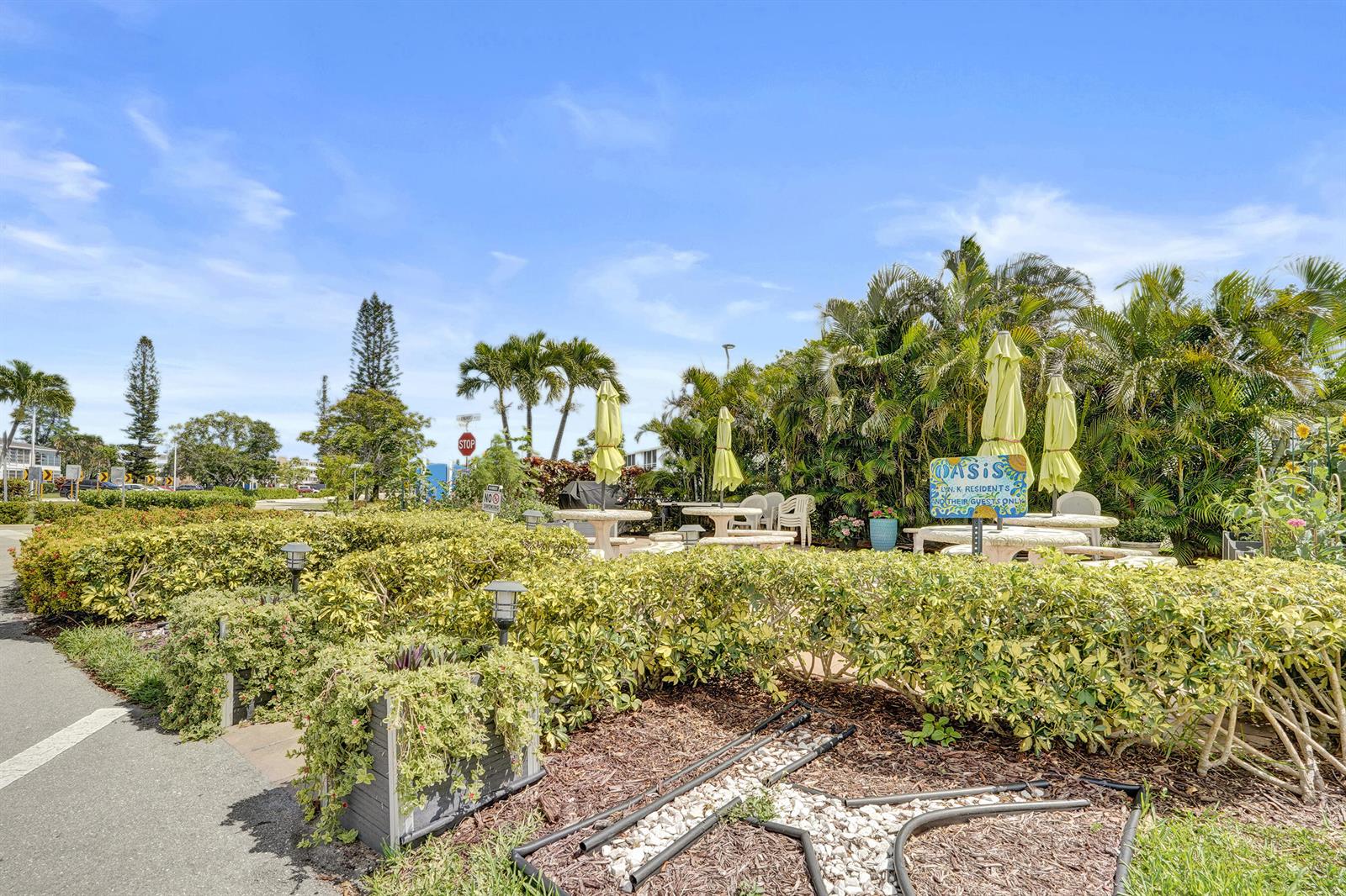 An outdoor patio area features tables with umbrellas, lush landscaping, and a sign for residents — 2049 Lyndhurst, Deerfield Beach FL 33442, DEERFIELD BEACH, FL