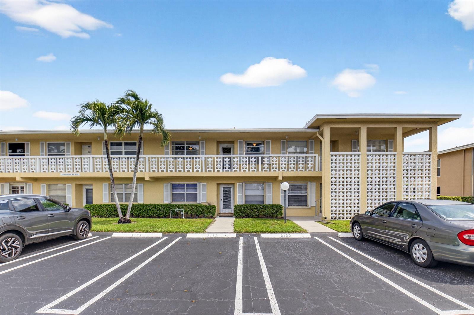 A two-story yellow apartment building features a parking lot and palm trees in front — 1150 Mahogany Way # 203, Delray Beach FL 33445, DELRAY BEACH, FL