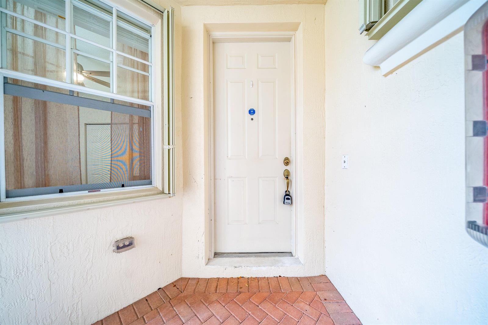 A white front door with a lockbox sits on a brick porch beside a window — 2106 Wingate Bend, Wellington FL 33414, WELLINGTON, FL