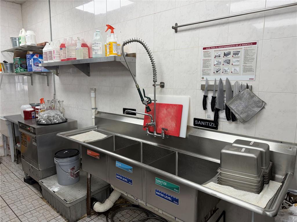 A commercial kitchen features a stainless steel three-compartment sink, wall-mounted faucet, and storage shelving — 9 NE PALM HARBOR VILLAGE WAY NE UNIT G, PALM COAST, FL