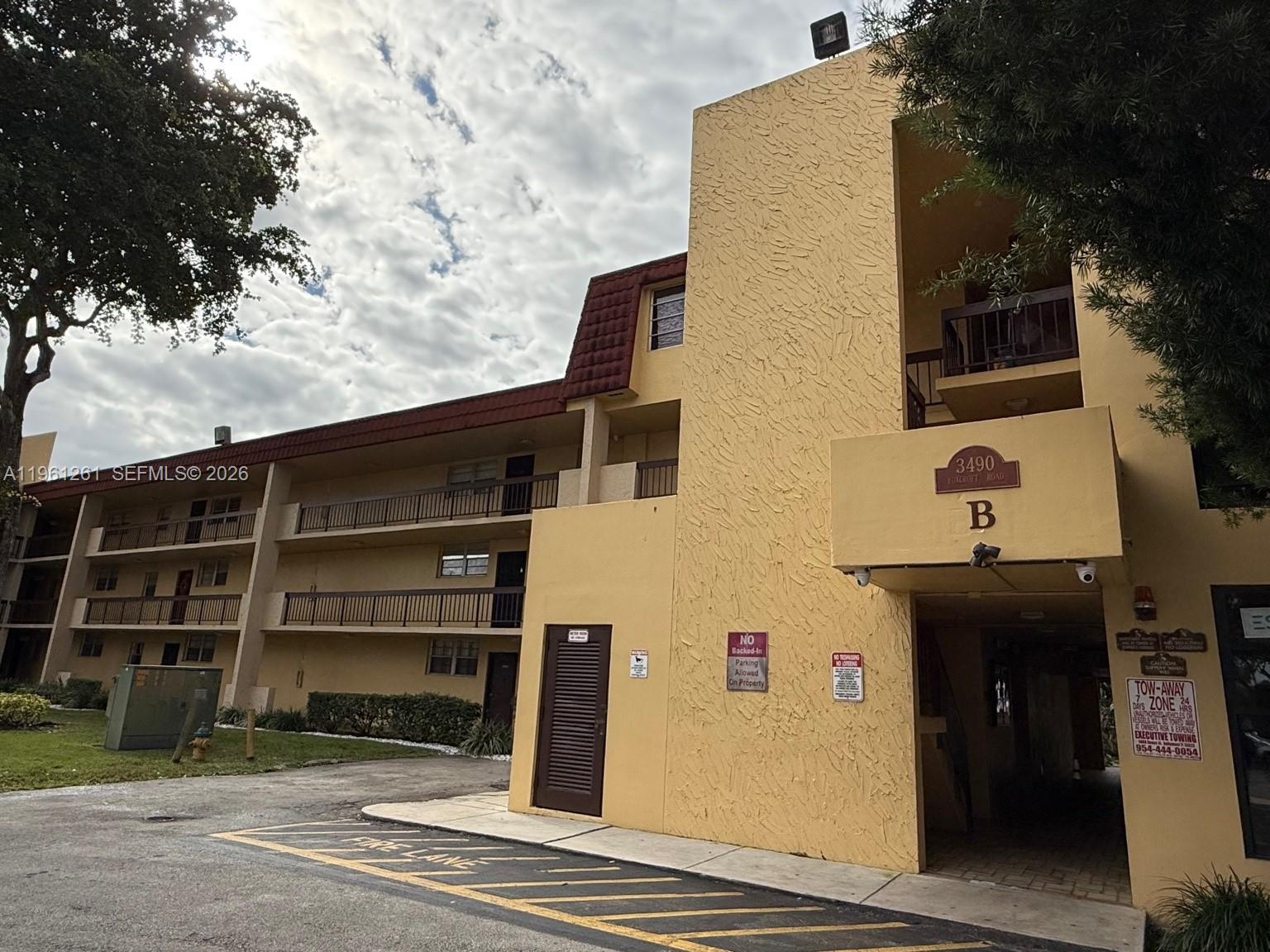 A three-story yellow apartment building with balconies and a covered entrance under a cloudy sky — 3490 Foxcroft Rd # B208, Miramar FL 33025, MIRAMAR, FL