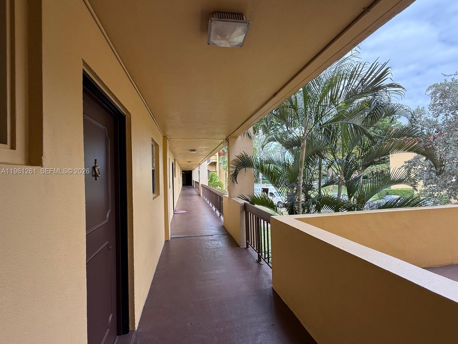 An outdoor apartment building walkway features a brown door, painted concrete floors, and railings — 3490 Foxcroft Rd # B208, Miramar FL 33025, MIRAMAR, FL