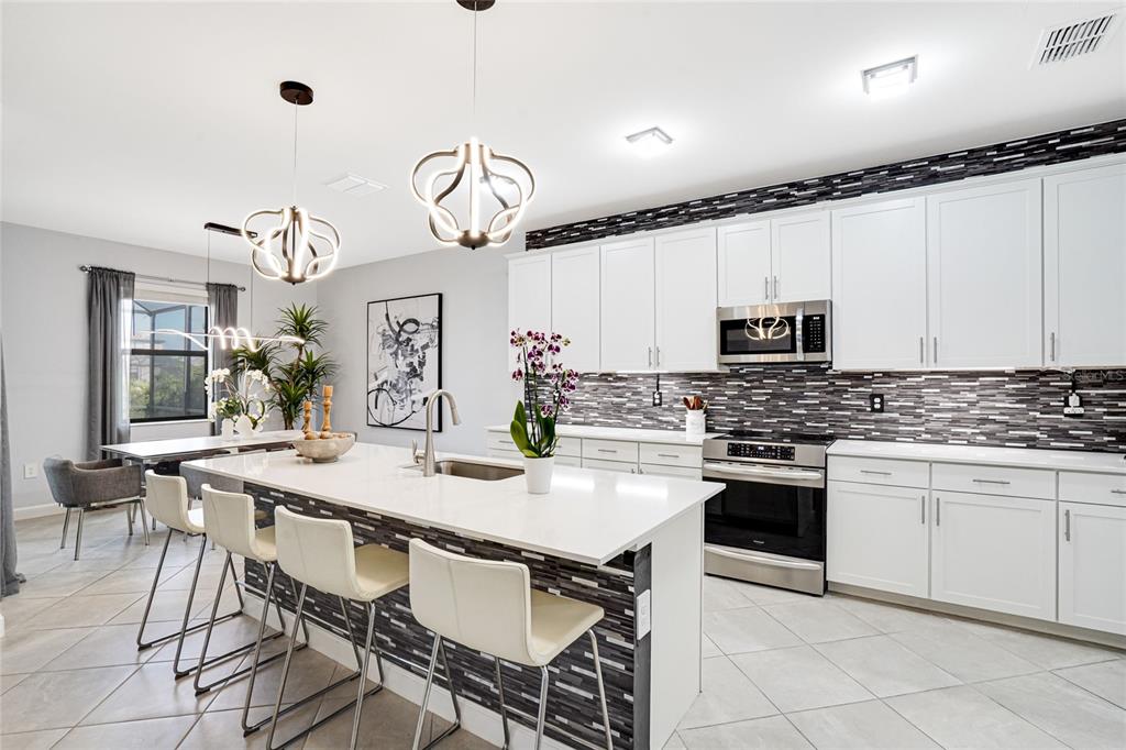 A modern kitchen features white cabinetry, a quartz island with bar seating, and tiled backsplash — 4970 OYSTER PEARL STREET, LAKEWOOD RANCH, FL