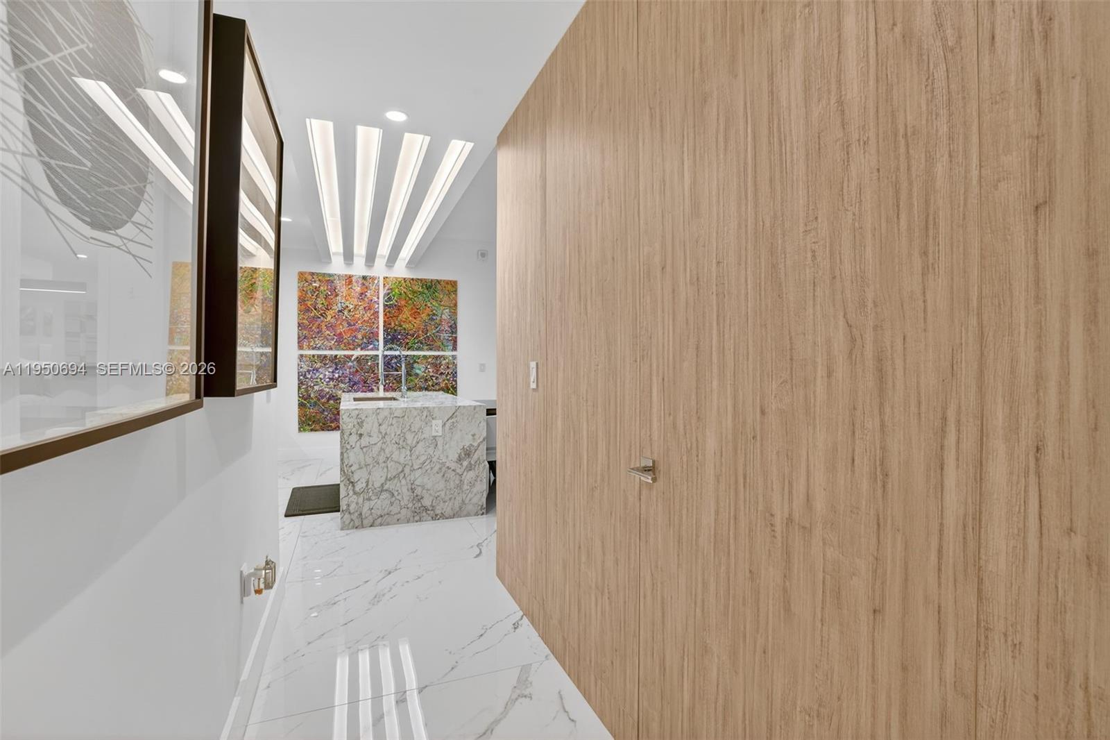 A hallway with wood-paneled walls leads to a kitchen area featuring marble counters and artwork — 5775 Collins Ave # 405, Miami Beach FL 33140, MIAMI BEACH, FL