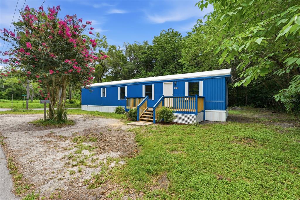 A blue mobile home with a wooden deck stands on a grassy lot with trees — 1212 NE 1ST AVENUE, WILLISTON, FL