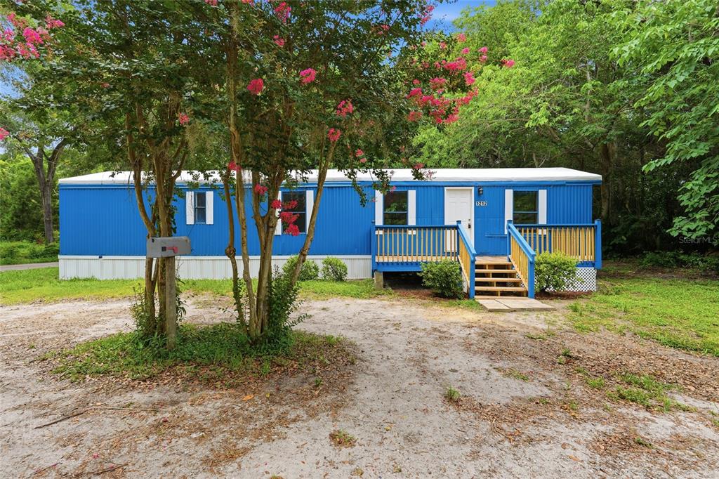 A blue mobile home with a wooden deck stands behind a flowering tree — 1212 NE 1ST AVENUE, WILLISTON, FL