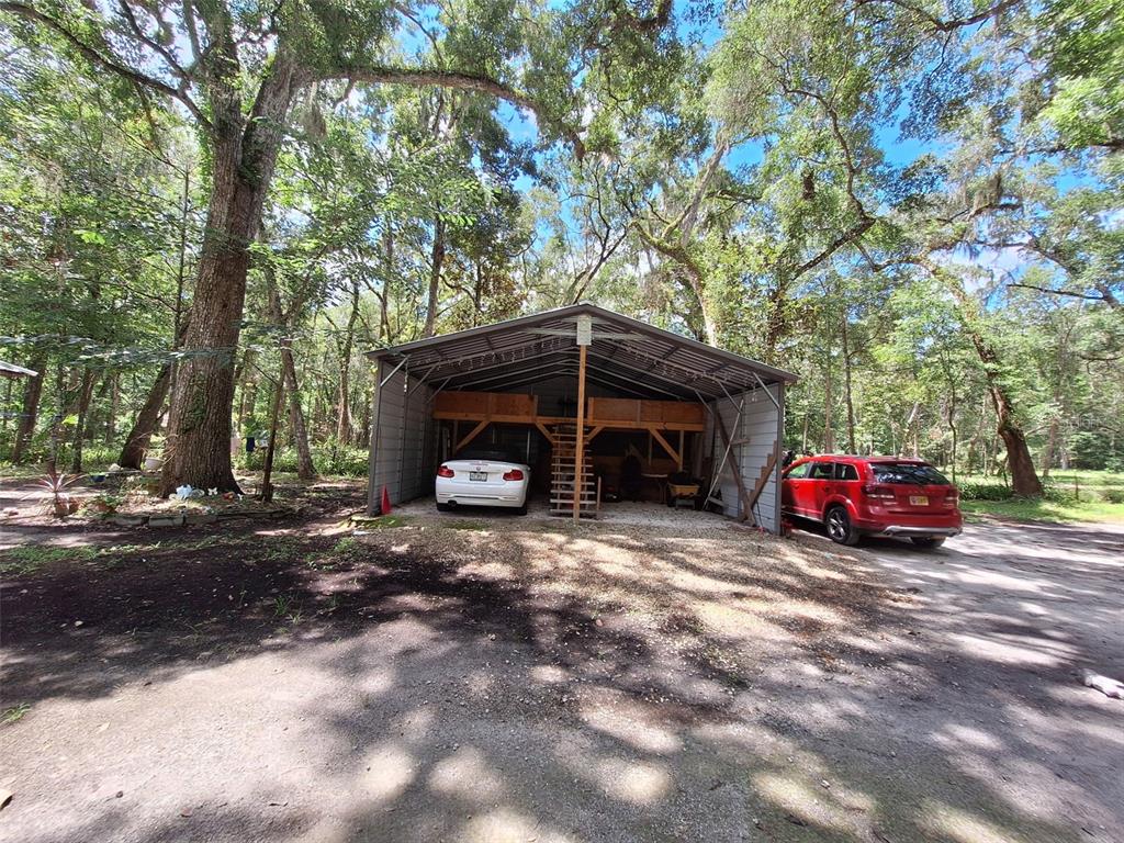 A metal carport with a wooden loft structure sits in a wooded area with two cars — 11304 ROYAL DRIVE, BROOKSVILLE, FL