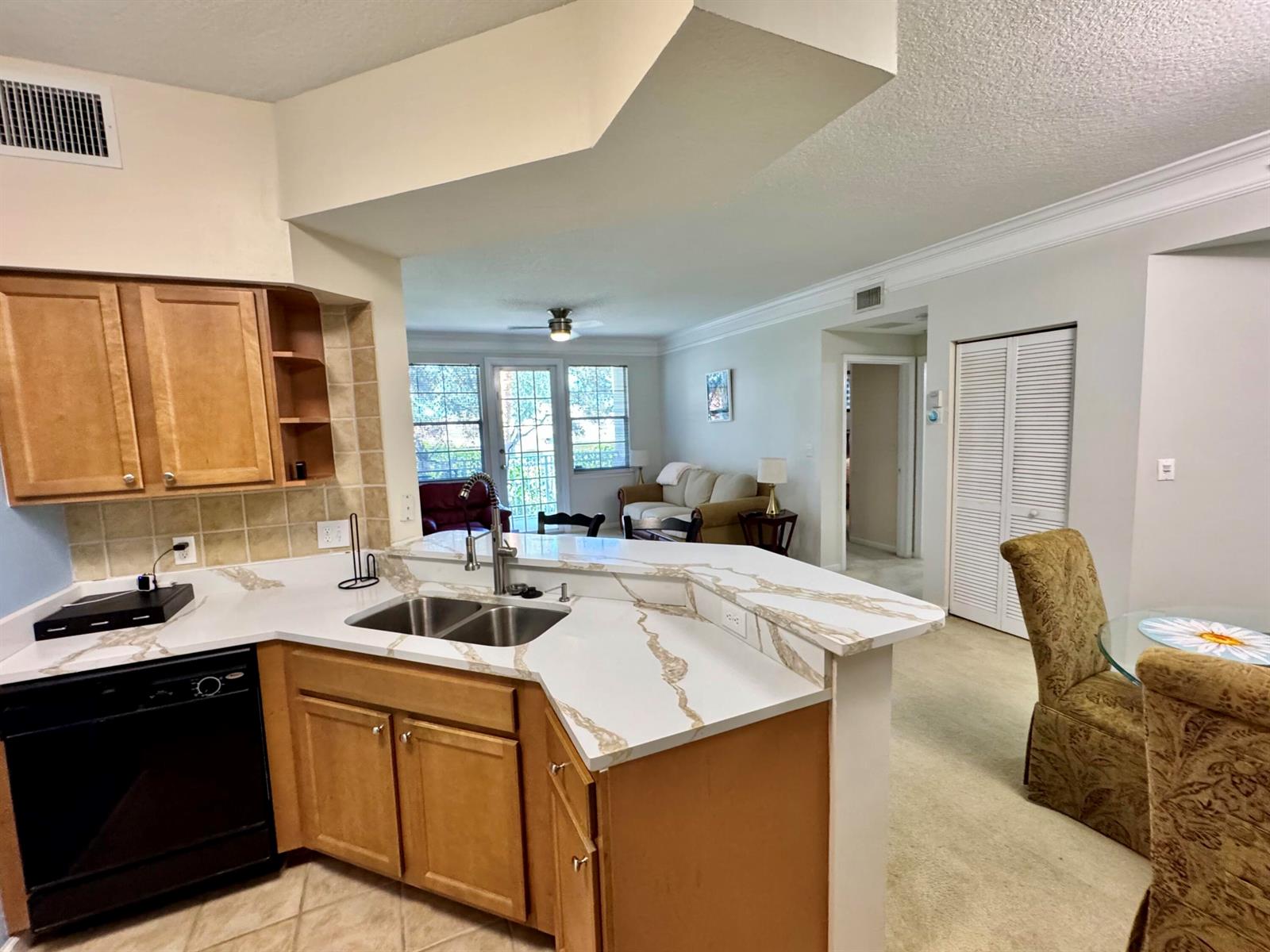A kitchen with light wood cabinets and stone countertops overlooks a living and dining area — 9857 Baywinds # 9210, West Palm Beach FL 33411, WEST PALM BEACH, FL