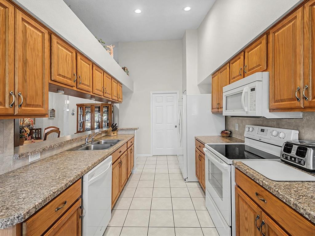 A galley kitchen features oak cabinets, granite-look countertops, white appliances, and light-colored tile flooring — 11625 BARONWOOD COURT, HUDSON, FL