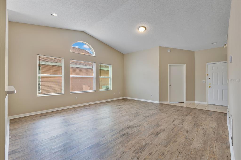 An empty living room features light wood-look flooring, vaulted ceilings, and large windows — 8821 PARLIAMENT COURT, KISSIMMEE, FL