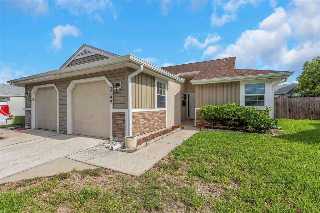 A single-story house with a two-car garage, tan siding, stone accents, and a front walkway — 3199 PHLOX DRIVE UNIT 214, PALM HARBOR, FL
