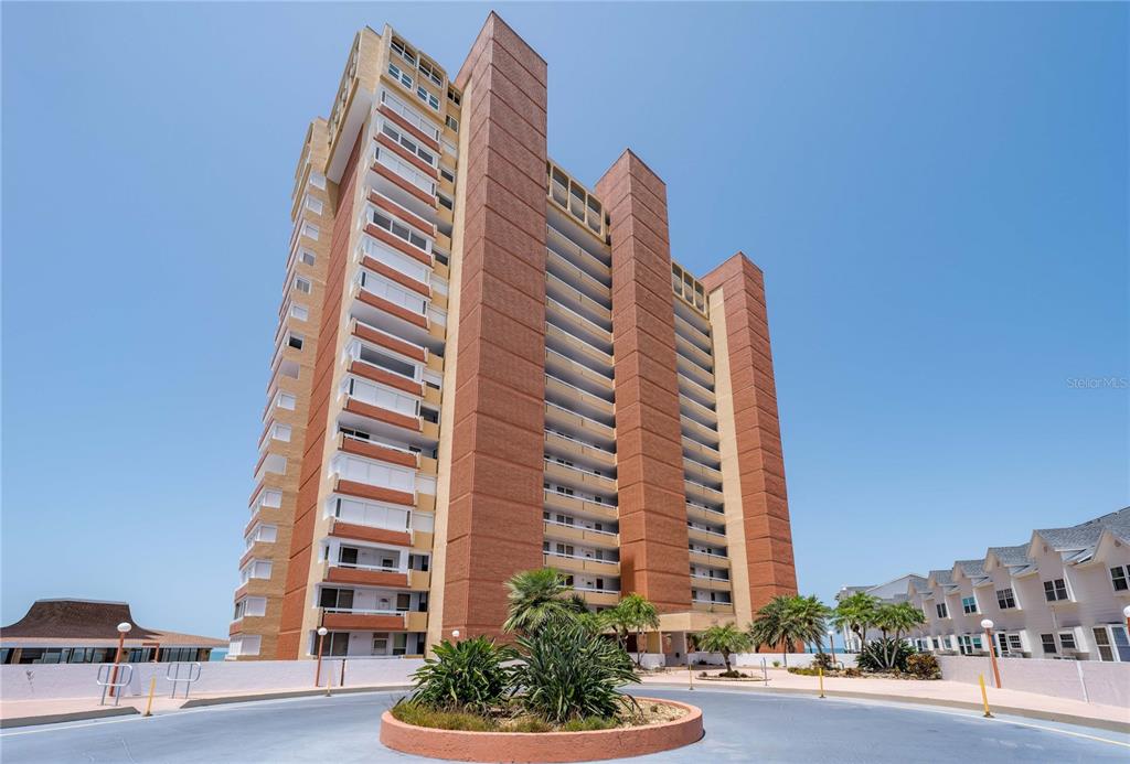 A tall brick high-rise apartment building stands behind a circular driveway with landscaping — 17940 GULF BOULEVARD UNIT 18C, REDINGTON SHORES, FL