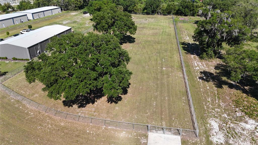 An aerial view shows a fenced, grassy lot with large oak trees next to warehouses — 980 DUCK BOX ROAD, UMATILLA, FL