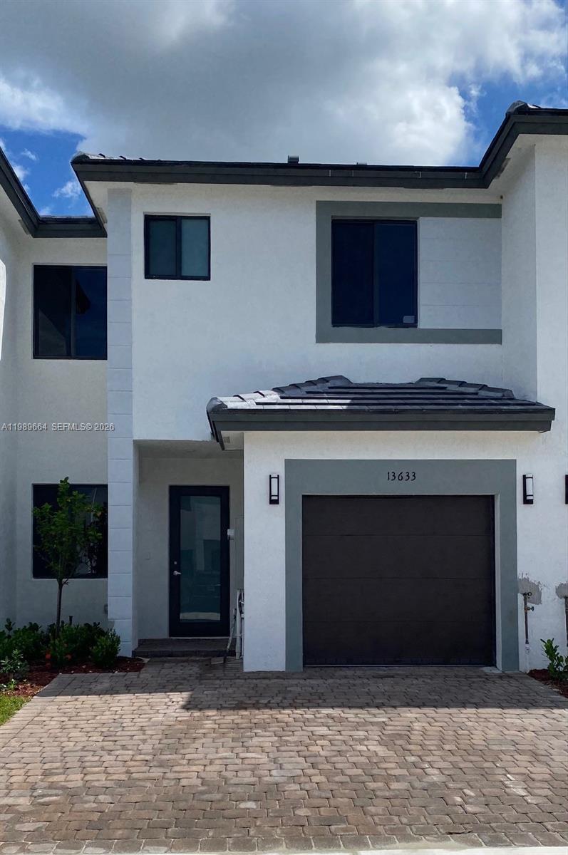 A two-story townhouse features a white stucco exterior, a dark garage door, and a paver driveway — 13633 SW 157th Pl # 0, Miami FL 33196, MIAMI, FL